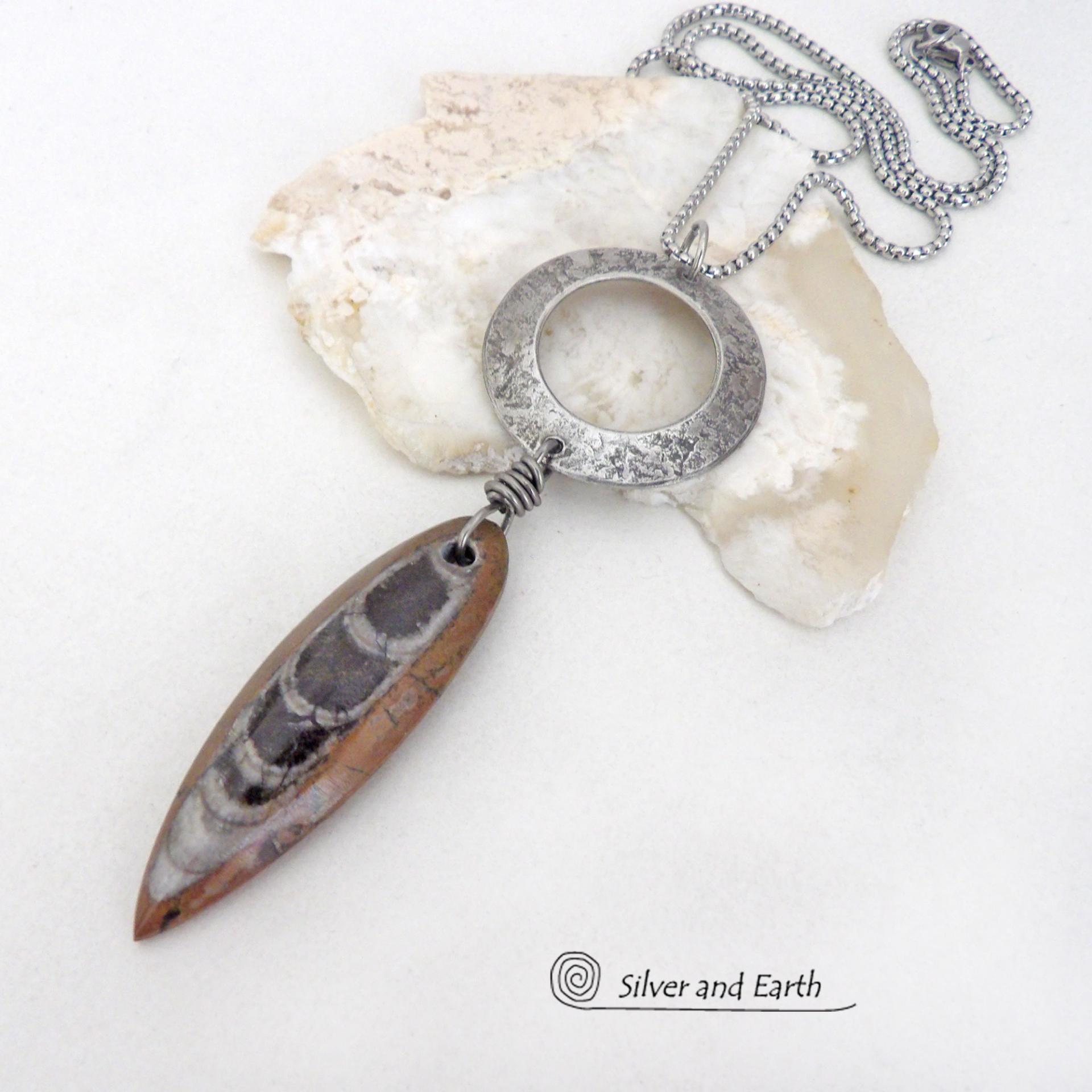 This one of a kind necklace features a natural Orthoceras fossil stone dangling from a silver pewter circle with a lightly hammered texture. This particular fossil has a brown hue - which is unusual, because these fossils are typically found in more black tones. This type of fossil is found in the Sahara Desert of Morocco where they were once ocean dwellers. Their shells were preserved in the marble in which they are found today. The design is unisex -- so it can be worn by either a man or a woman. Makes a 