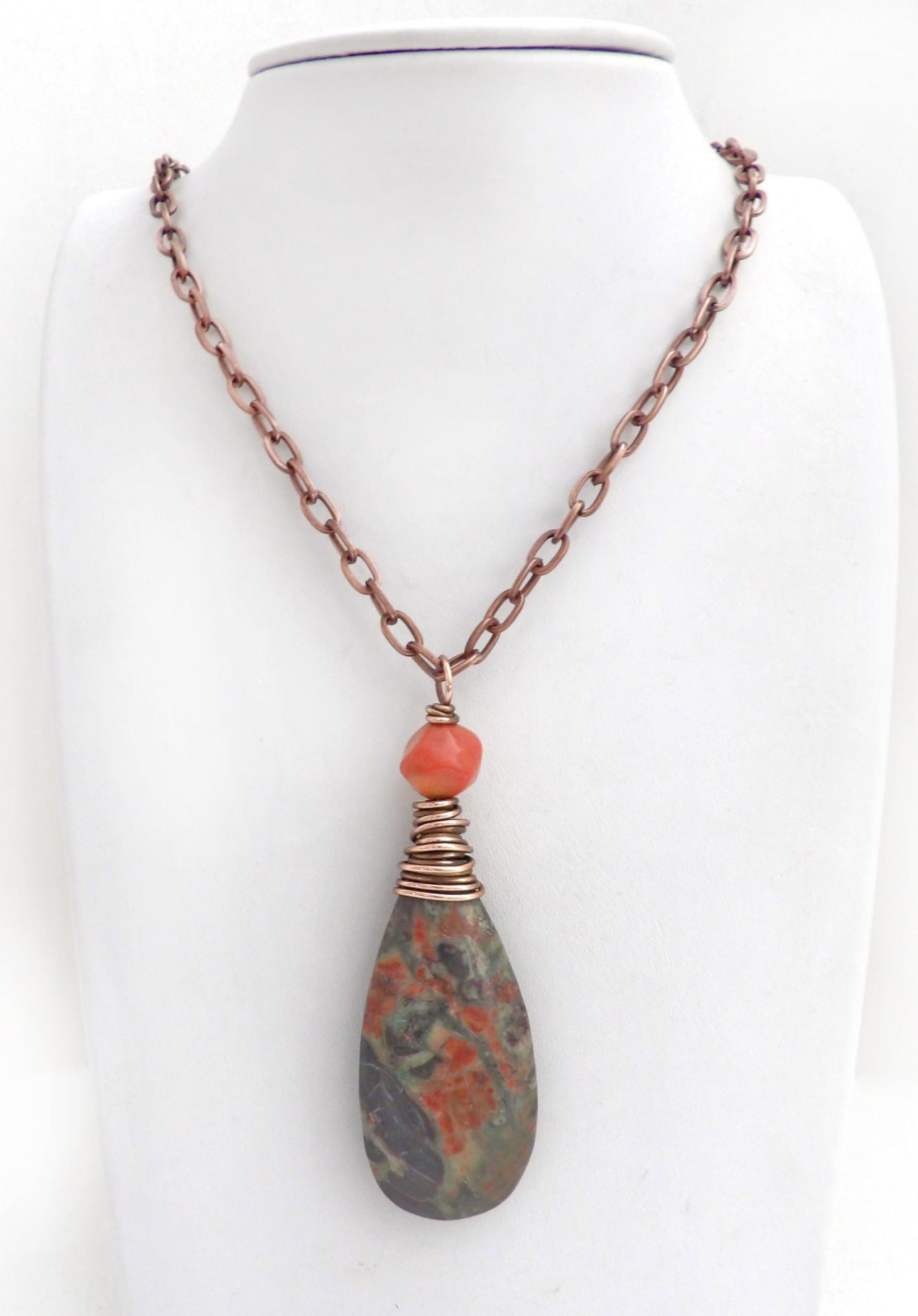 This handcrafted pendant necklace features a natural rhyolite jasper stone wire wrapped in copper wire and accented with a nugget-shaped coral bead. It has a natural pattern in several shades of earthy green along with splashes of orange. It has an earthy, casual boho style and is perfect for anyone that loves natural stones. It comes on an 18” copper chain with sturdy hook & eye closure; which can be removed from the pendant and worn alone or with other pendants in your wardrobe.