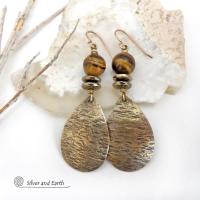 These handcrafted earrings feature textured gold brass teardrop dangles accented with natural brown tiger's eye gemstones and hematite stone beads. The beads have an electroplated champagne gold tone finish which adds a touch of shimmer and shine. They have an earthy modern style that coordinates well with all wardrobe styles. Handmade in the USA by Silver and Earth Jewelry.