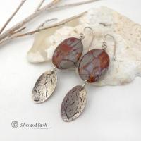 These handcrafted one-of-a-kind earrings feature natural Noreena jasper gemstones accented with textured sterling silver dangles. This stone is known for its dramatic abstract and geometric patterns and is found only in the Pilbara region of Western Australia. The stones in this pair have a great pattern consisting of hues of deep burgundy-red with fiery orange and gray streaks. They'll make a great gift for women that appreciate and love unique natural stones. 