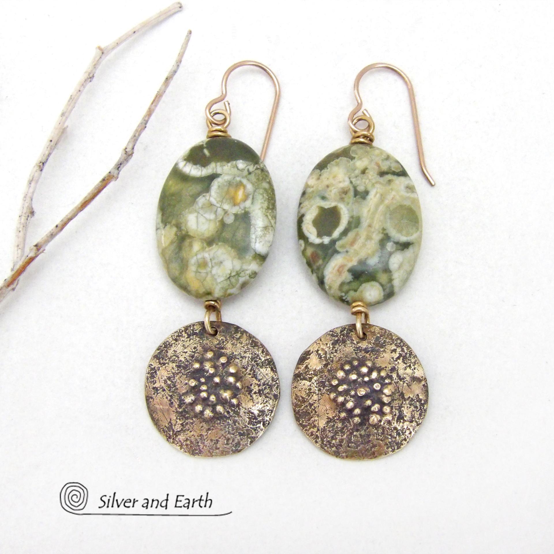 These earrings feature gold brass dangles with a rustic hammered and hand stamped texture accented with natural green rainforest jasper gemstones. They have an earthy chic, modern style that coordinates well with all wardrobe types. Rainforest Jasper is a gemstone that belongs to the rhyolite family. It's known for its vibrant patterns and earthy tones featuring a mix of greens, browns, and cream. These colors form beautiful and intricate patterns which resemble the lush, diverse vegetation of a tropical ra