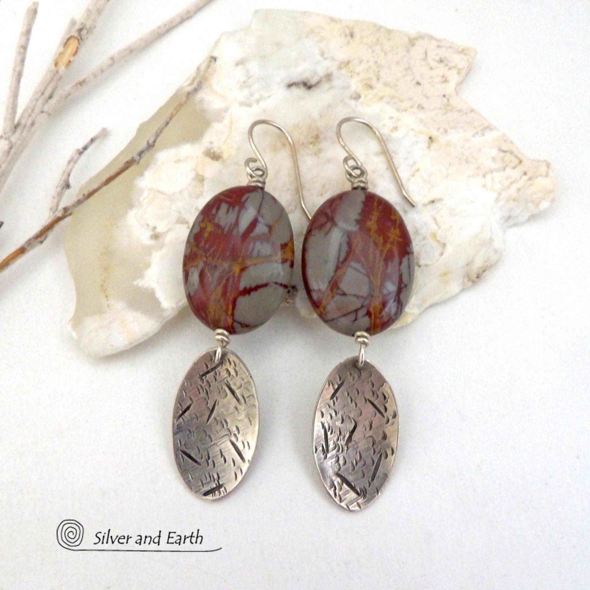 These handcrafted one-of-a-kind earrings feature natural Noreena jasper gemstones accented with textured sterling silver dangles. This stone is known for its dramatic abstract and geometric patterns and is found only in the Pilbara region of Western Australia. The stones in this pair have a great pattern consisting of hues of deep burgundy-red with fiery orange and gray streaks. They'll make a great gift for women that appreciate and love unique natural stones. 