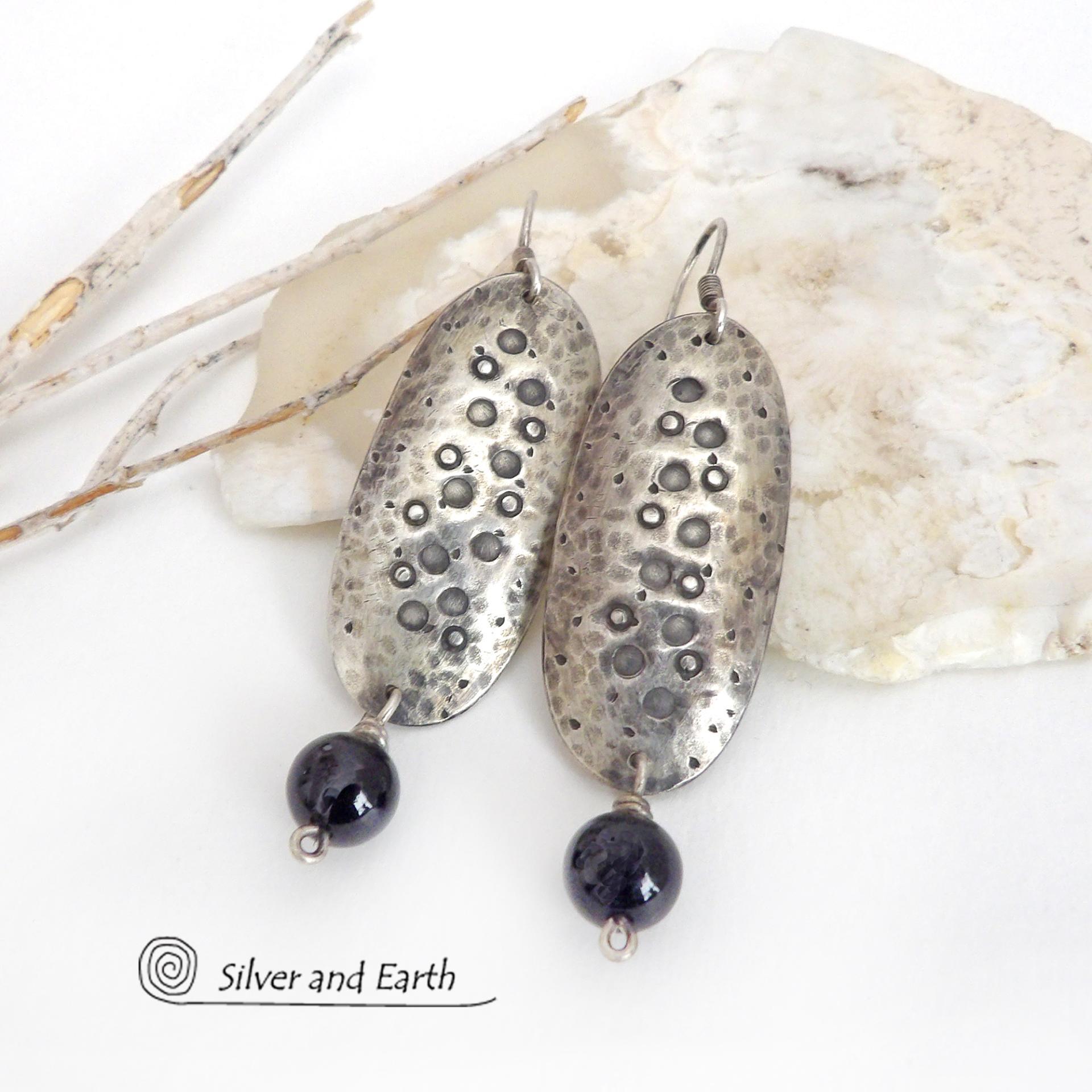 These handcrafted sterling silver earrings have a long oval shape with a hammered and hand stamped texture. Black onyx gemstones dangle from the bottom. They have a modern, earthy organic vibe that coordinates well with all wardrobe styles. Earrings are handmade in the USA by Silver and Earth Jewelry. 