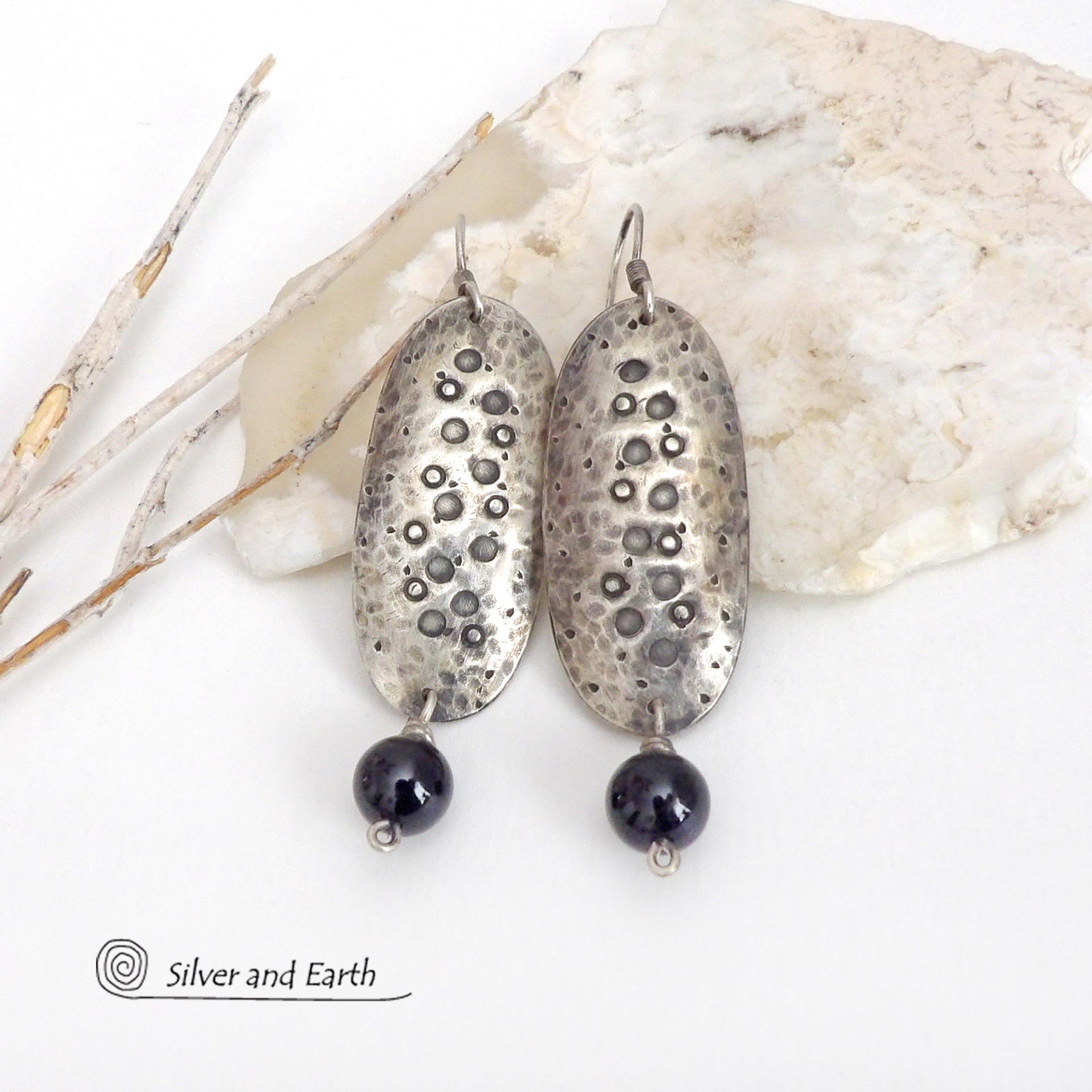 These handcrafted sterling silver earrings have a long oval shape with a hammered and hand stamped texture. Black onyx gemstones dangle from the bottom. They have a modern, earthy organic vibe that coordinates well with all wardrobe styles. Earrings are handmade in the USA by Silver and Earth Jewelry. 
